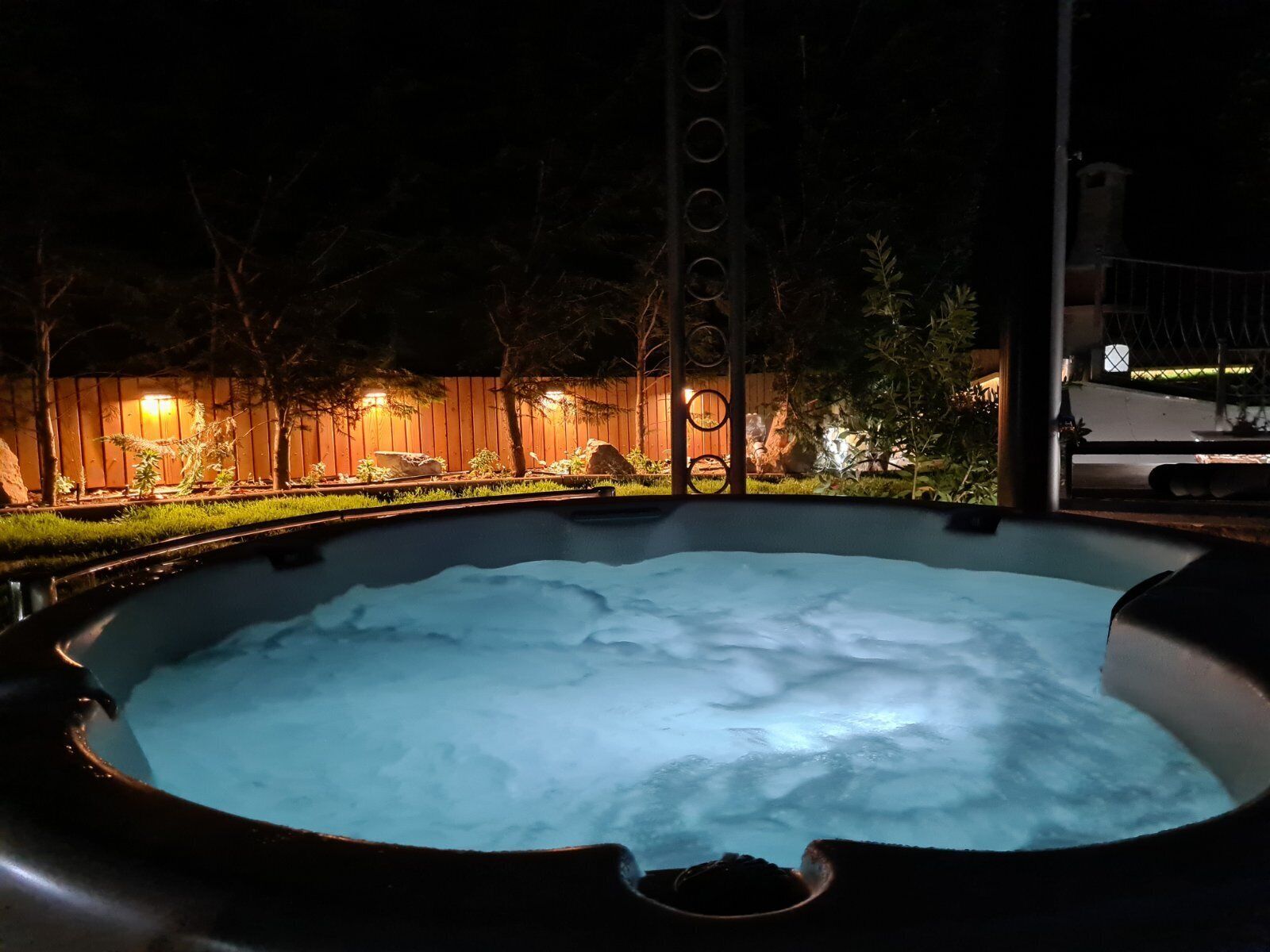Een hottub omringd door een houten omheining en een kleine tuin met planten en een boom.