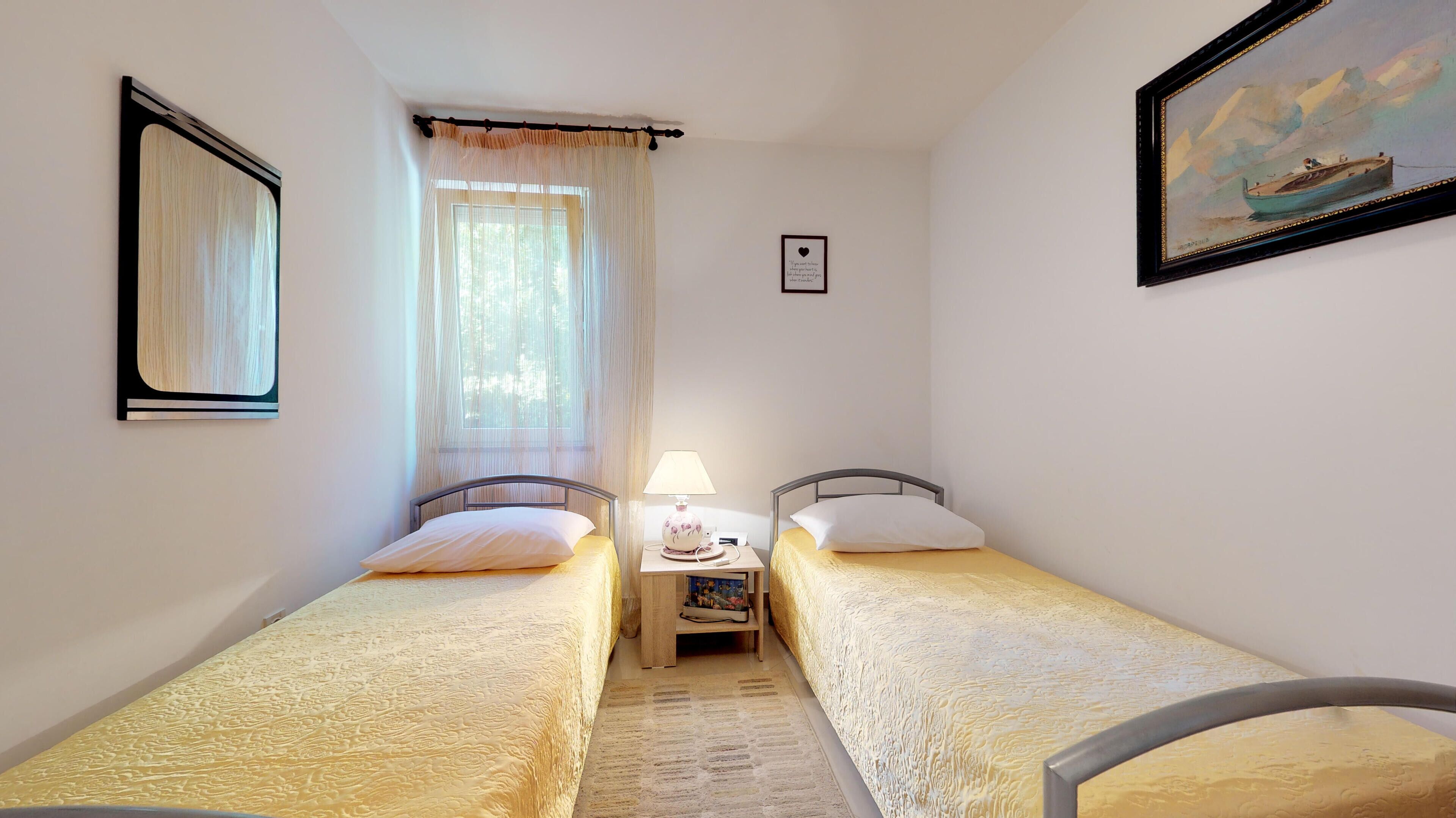 Een kleine, schone kamer met twee eenpersoonsbedden, een nachtkastje met een lamp, een raam met dunne gordijnen en een lijst met een afbeelding aan de muur.