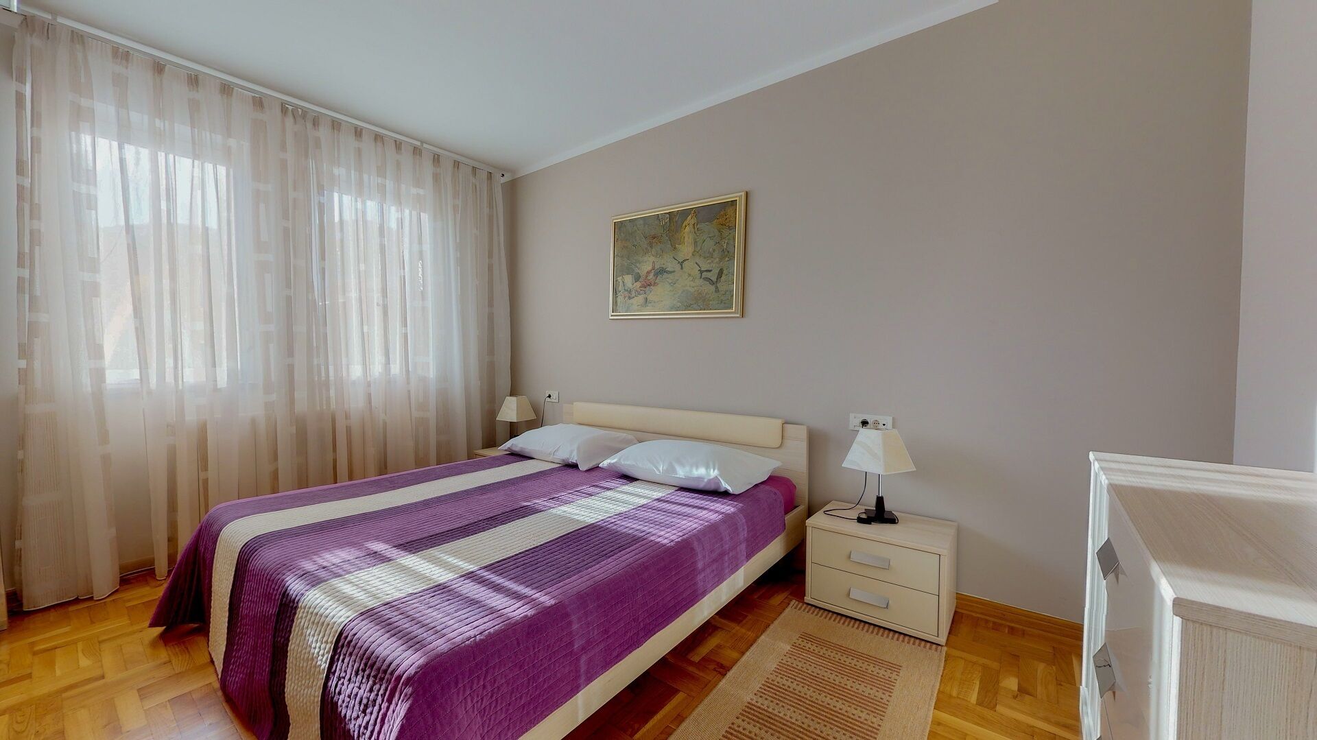 A bedroom with a bed, a nightstand, a lamp, a painting on the wall, and a window with sheer curtains.