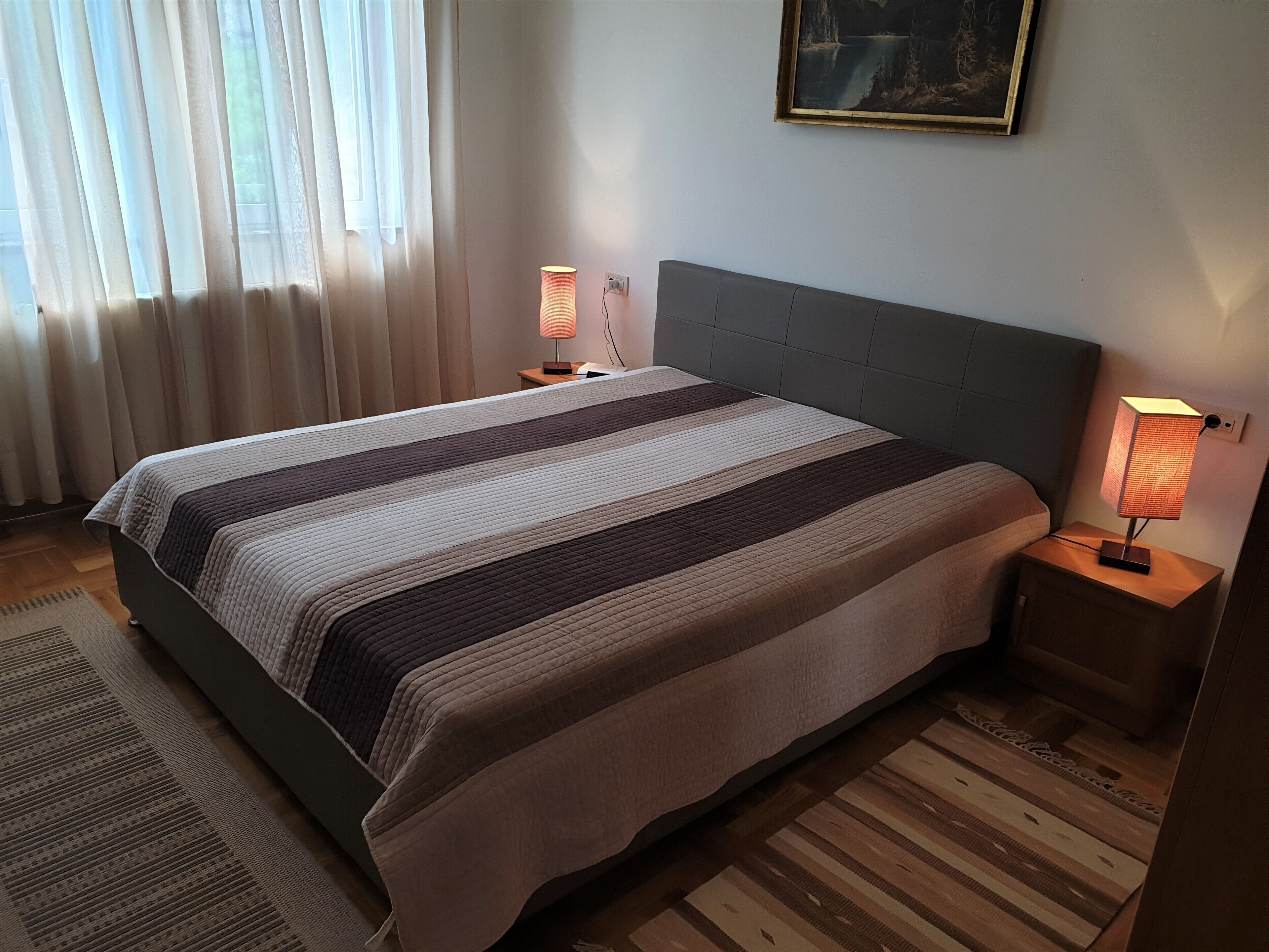 A neatly made bed with a striped blanket, two bedside lamps, a painting on the wall, and a window with curtains.
