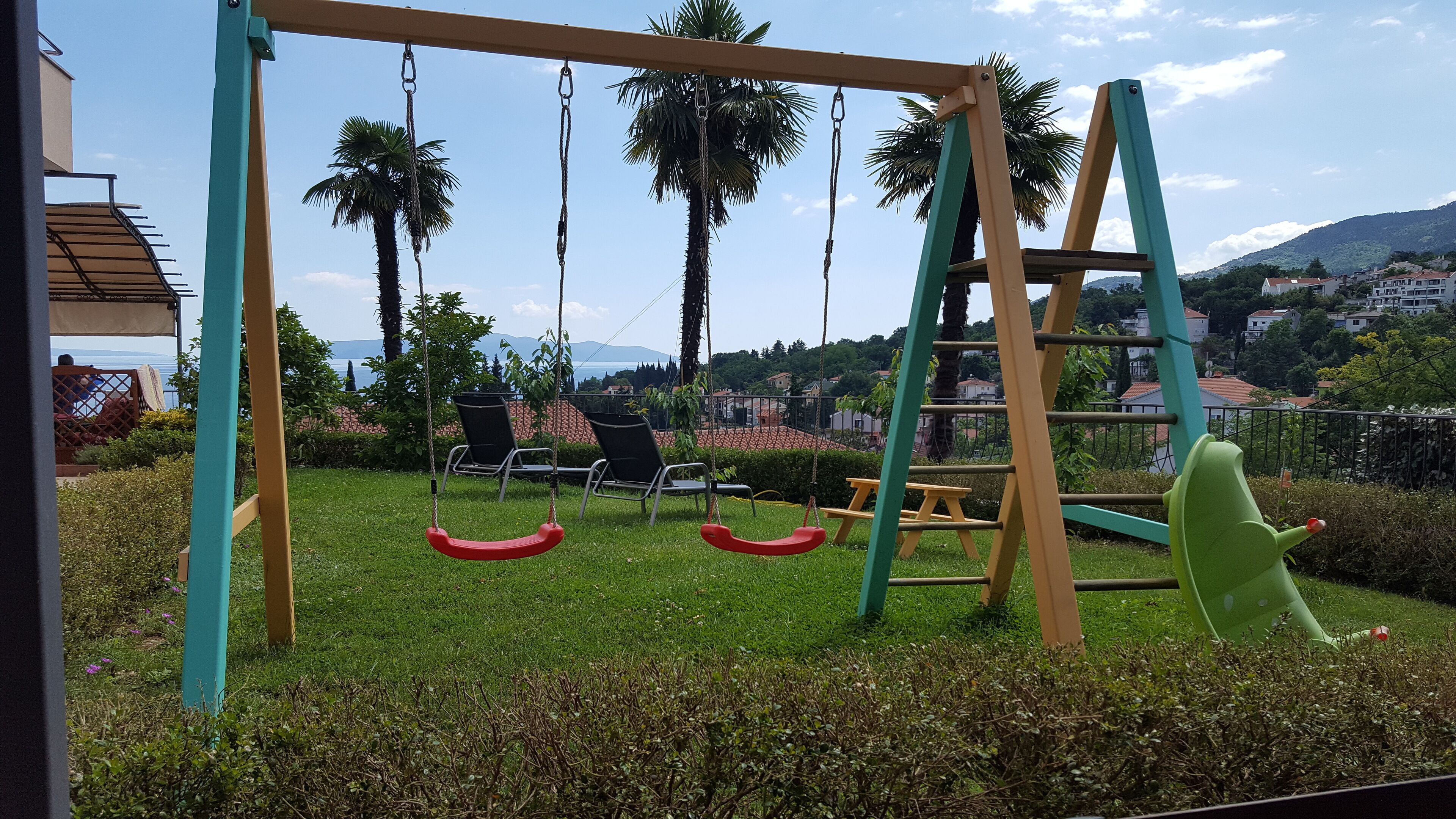 Un parc infantil amb cadira d'oscil·lació i tobogan, envoltat de vegetació i palmeres, amb vista a cases i muntanyes al fons.