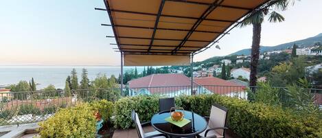 Eine Terrasse mit Tisch und Stühlen, Blick auf das Meer und eine Stadt im Hintergrund.
