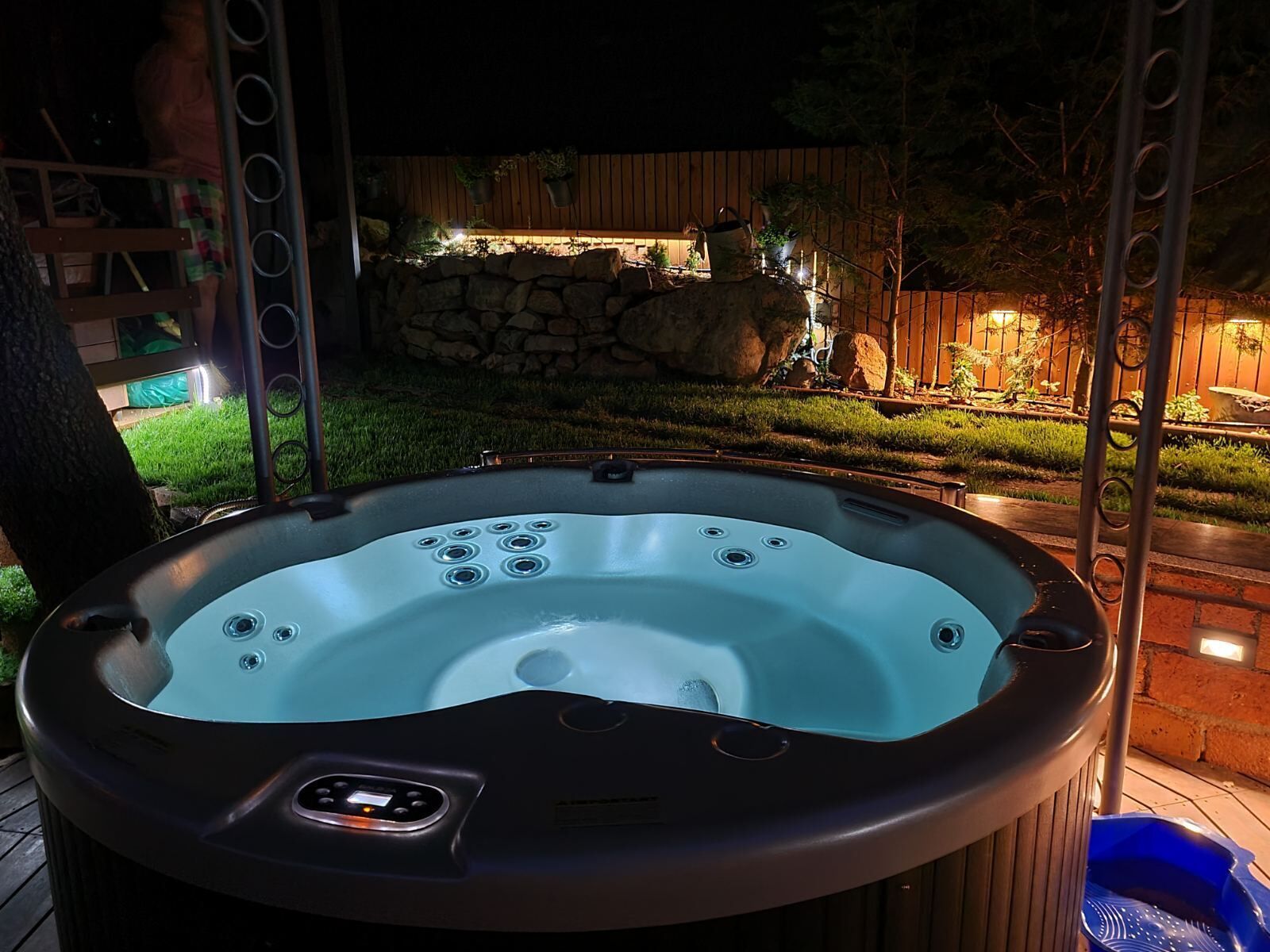 Un bain à remous doté d’un panneau de commande, entouré d’un mur de pierres, d’une clôture en bois et d’un espace jardin bien éclairé la nuit.
