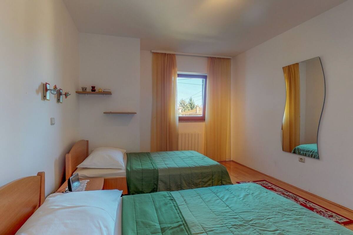 Une chambre d’hôtel avec deux lits simples, une fenêtre avec des rideaux, un miroir fixé au mur et une petite étagère avec des objets décoratifs.