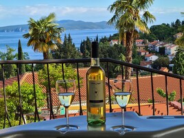 Un balcón con una botella de vino y dos copas, con vistas a un pueblo costero con casas y al mar.