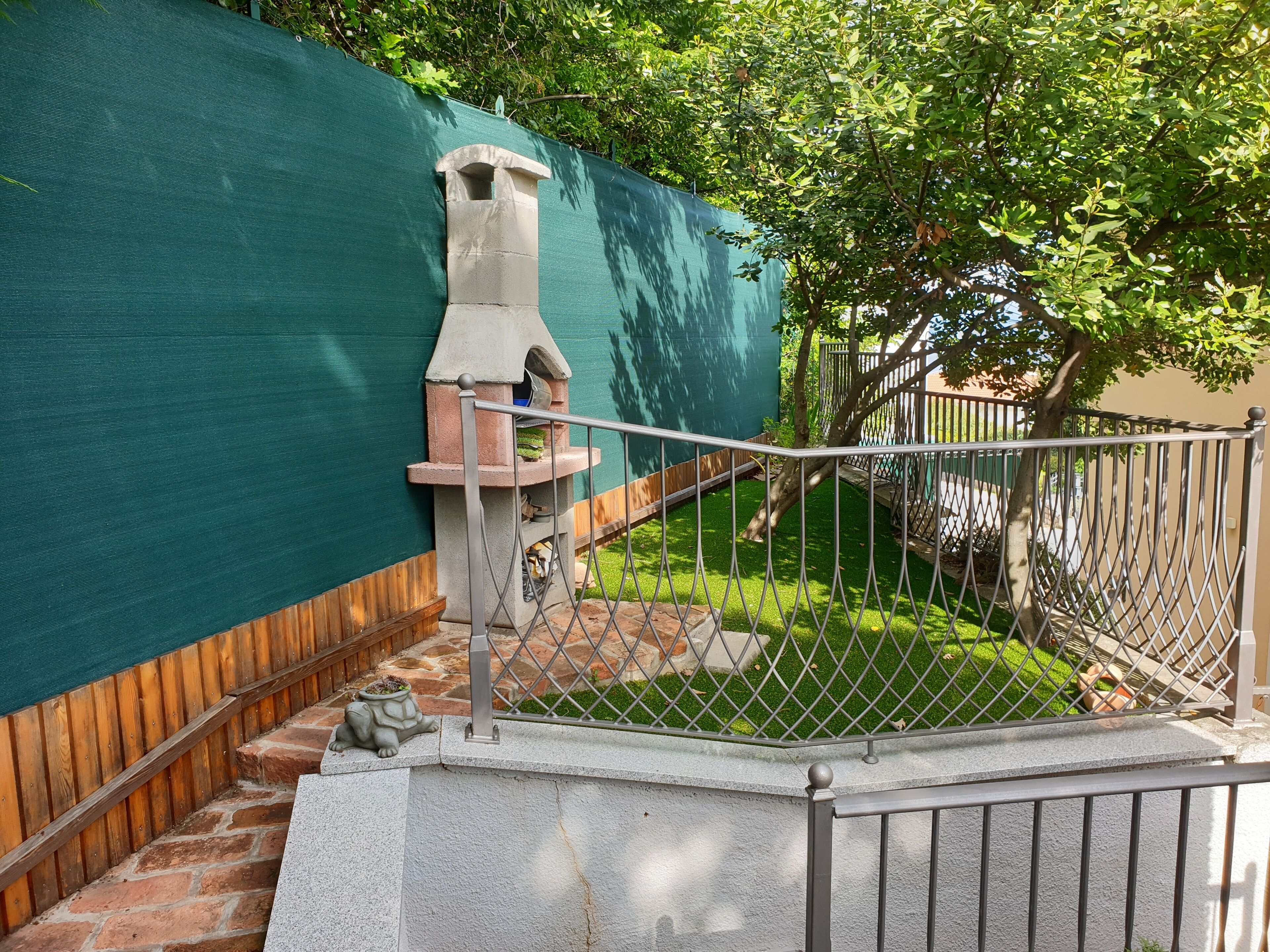 Un patio trasero con una parrilla de piedra, una valla de madera y un muro verde.