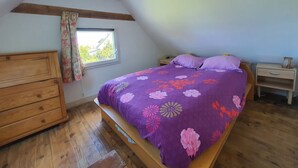 A bedroom with a wooden bed, a nightstand, a dresser, and a window with curtains. - Petite Maison Proche Mer, Jardin Clos, Près du GR 34 et du Bourg , Chien Accepté (Plouguerneau)