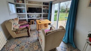 A living room with patterned floor tiles, a sofa, a chair, a wooden table, and a bookshelf. - Petite Maison Proche Mer, Jardin Clos, Près du GR 34 et du Bourg , Chien Accepté (Plouguerneau)