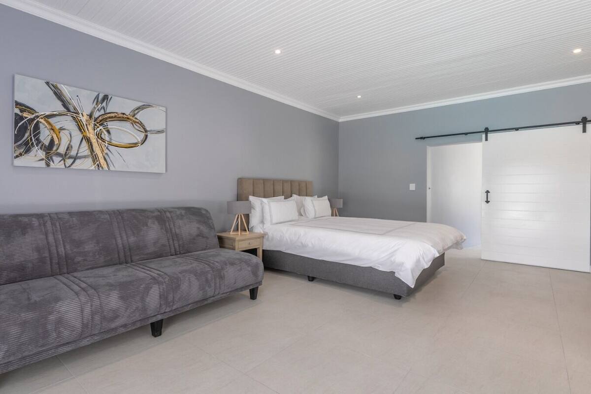 A modern bedroom with a grey sofa, a bed with white bedding, a nightstand, and a wall-mounted abstract painting.