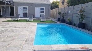 A rectangular swimming pool with a stone tile deck, two lounge chairs, and a small grassy area with a building in the background. - Inkantu Apartments & Suites (Cape Town)