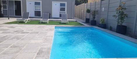 Ein rechteckiger Pool mit Steinplatten umrandet, zwei Liegestühle und eine kleine Rasenfläche mit einem Gebäude im Hintergrund.