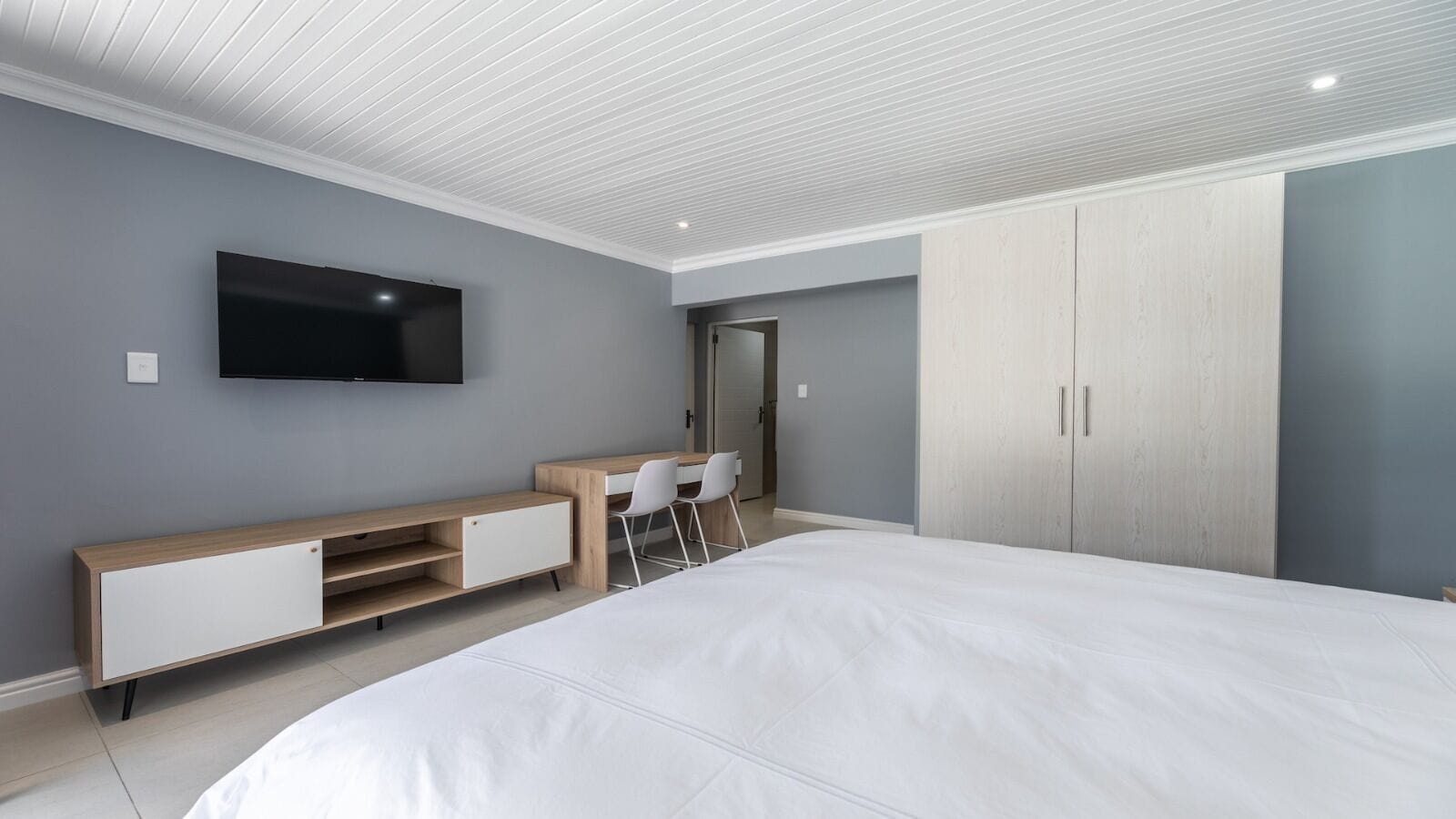 A modern bedroom with a large bed, a TV mounted on the wall, a desk with chairs, and a wardrobe.