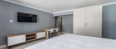 A modern bedroom with a large bed, a TV mounted on the wall, a desk with chairs, and a wardrobe.