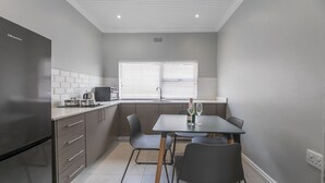 A compact kitchen with a refrigerator, stove, and sink, a dining table with chairs, and a window with blinds. - Inkantu Apartments & Suites (Cape Town)