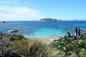 Un paysage côtier avec un phare situé sur une petite île, des eaux bleues et limpides, et une plage de sable bordée de végétation.