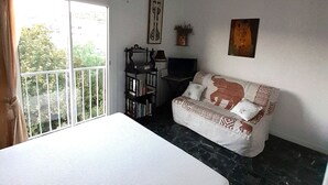 A bedroom with a bed, a sofa, a TV, a bookshelf, and a balcony with a view of greenery.