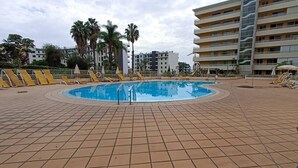 Ein Pool mit Fliesenboden, umgeben von Liegestühlen und Palmen, mit mehrstöckigen Gebäuden im Hintergrund.