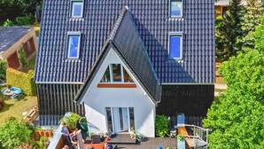 Ein zweistöckiges Haus mit einem steilen Satteldach, einer kleinen angebauten Garage und einer Veranda mit Sitzgelegenheiten.