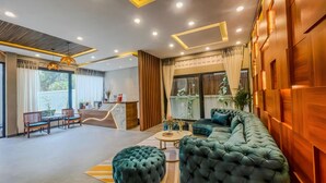 A modern living room with a sofa, ottomans, and a coffee table. There is a reception desk with a computer and a chair. The room has wooden paneling and a large window with curtains. - The Luxury (Siem Reap)