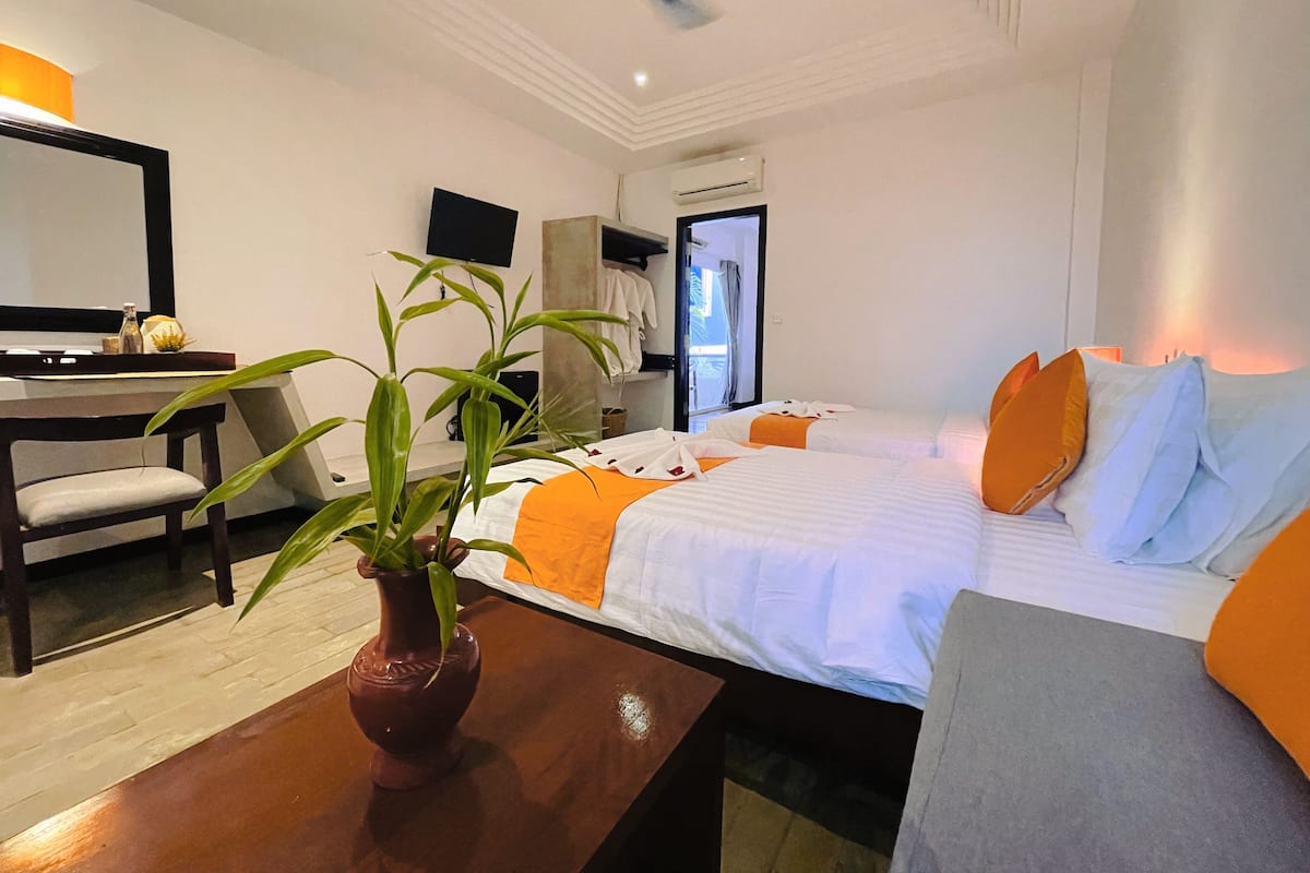 A hotel room with a bed, a desk, a chair, a plant, and an air conditioning unit.