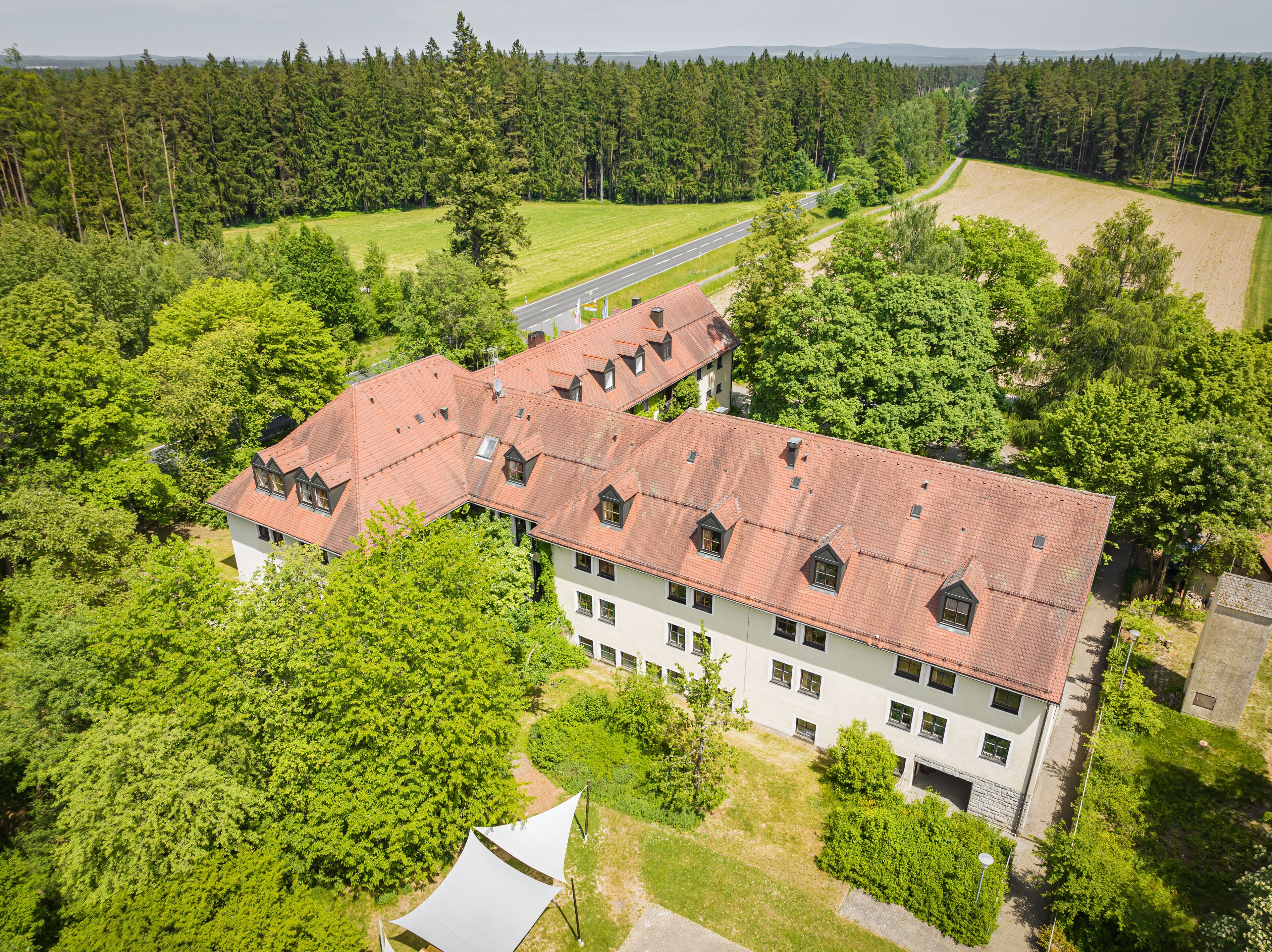 A large building with multiple floors and a red-tiled roof, surrounded by lush greenery and a road.