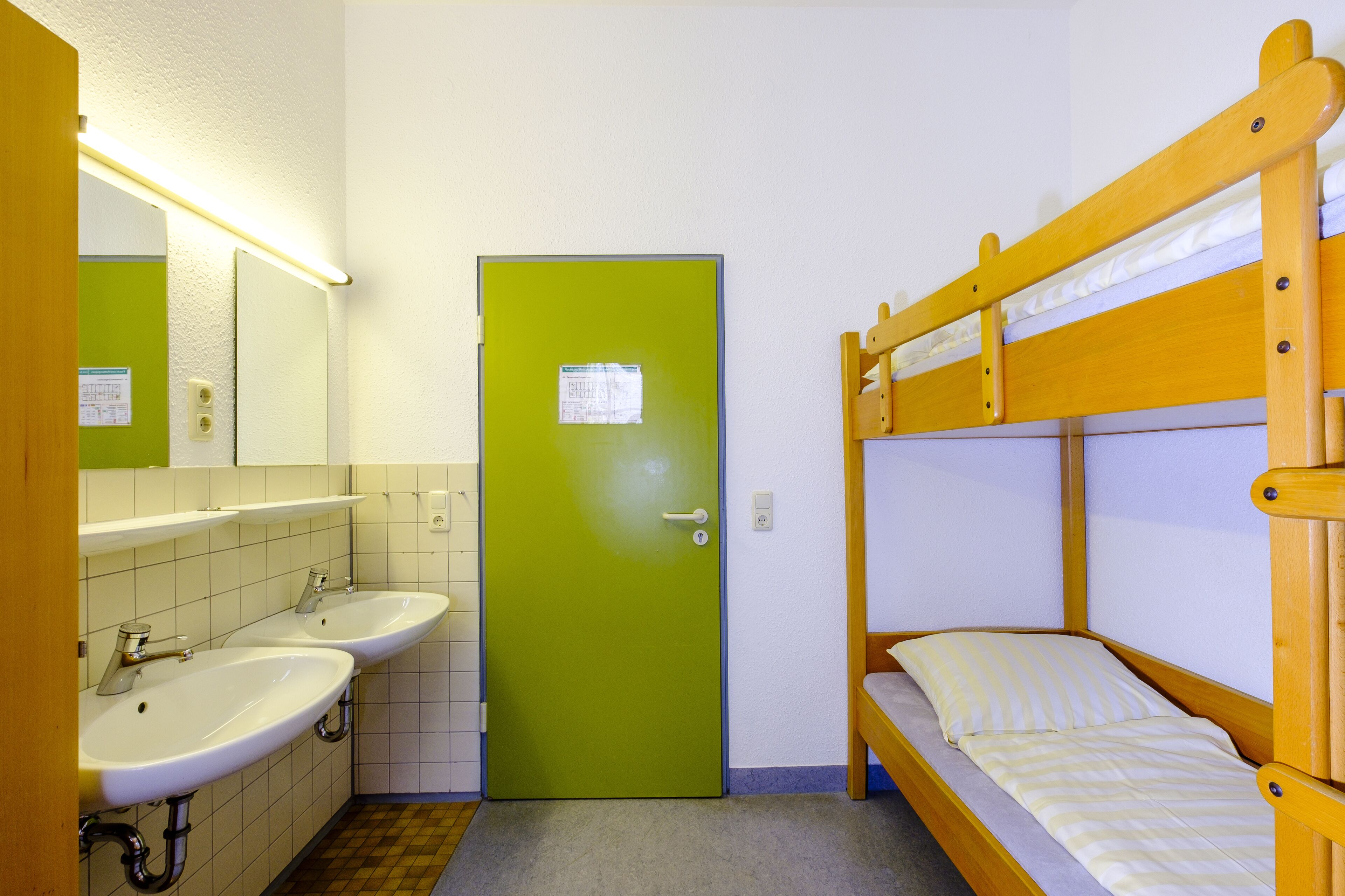 A small, single-room hotel bathroom with two sinks, a green door, and a wooden bunk bed.