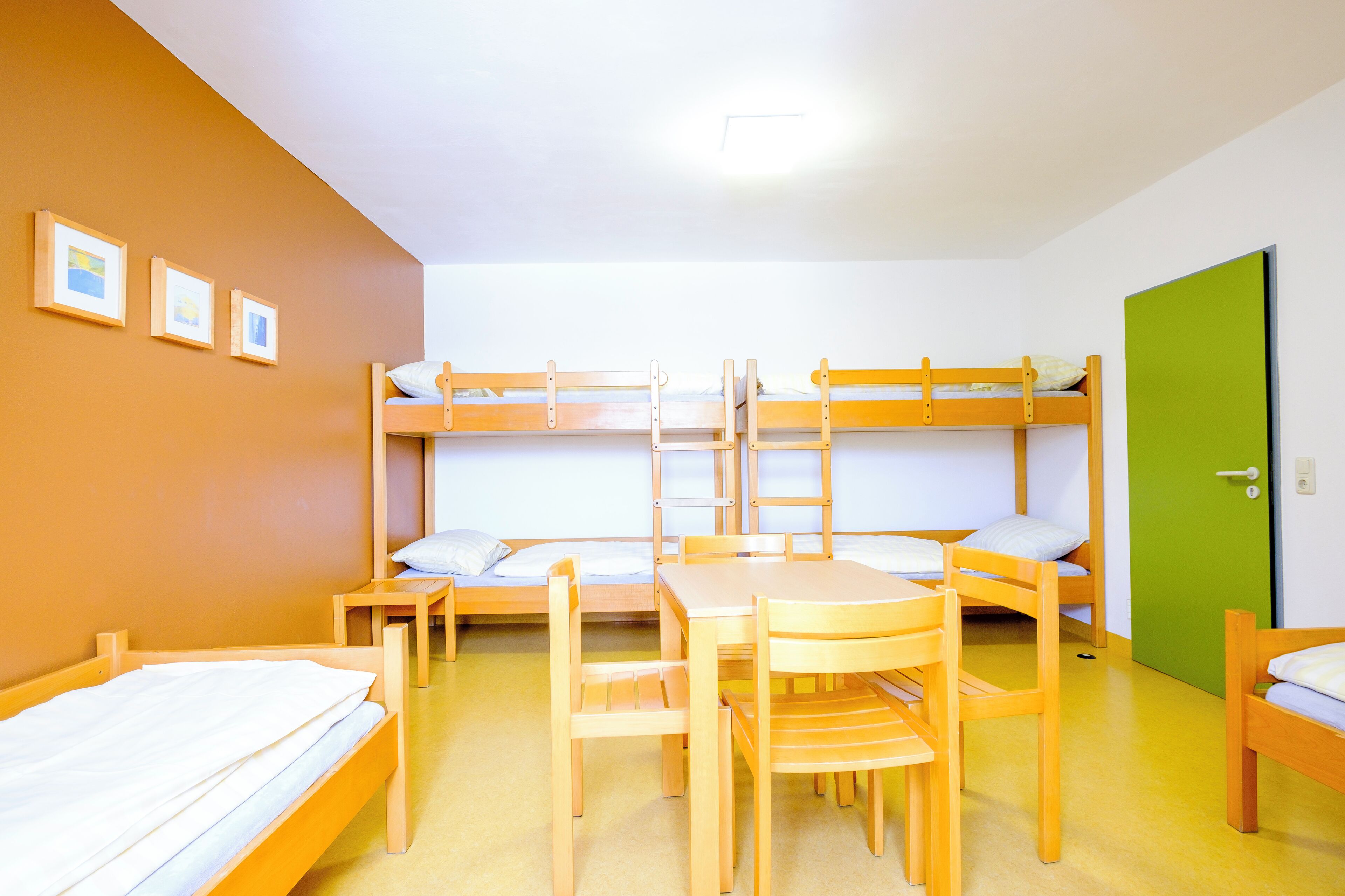 A dormitory room with bunk beds, a wooden table, chairs, and a green door.