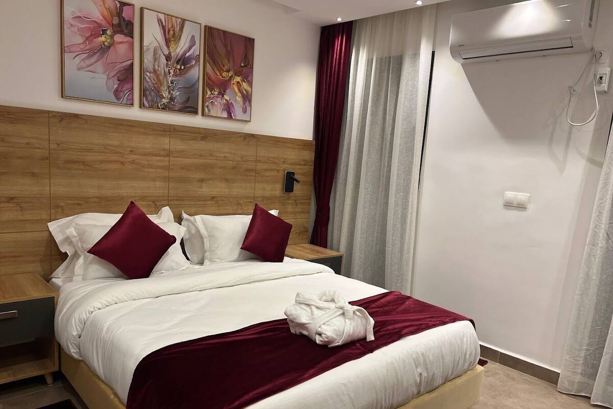 Cama bem arrumada com roupa de cama branca e bordô, cabeceira de madeira e manta vermelha. Há dois interruptores de luz na parede e um aparelho de ar condicionado branco.