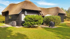 Una casa con techo de paja, un pequeño porche, una parrilla cubierta y una silla de jardín.
