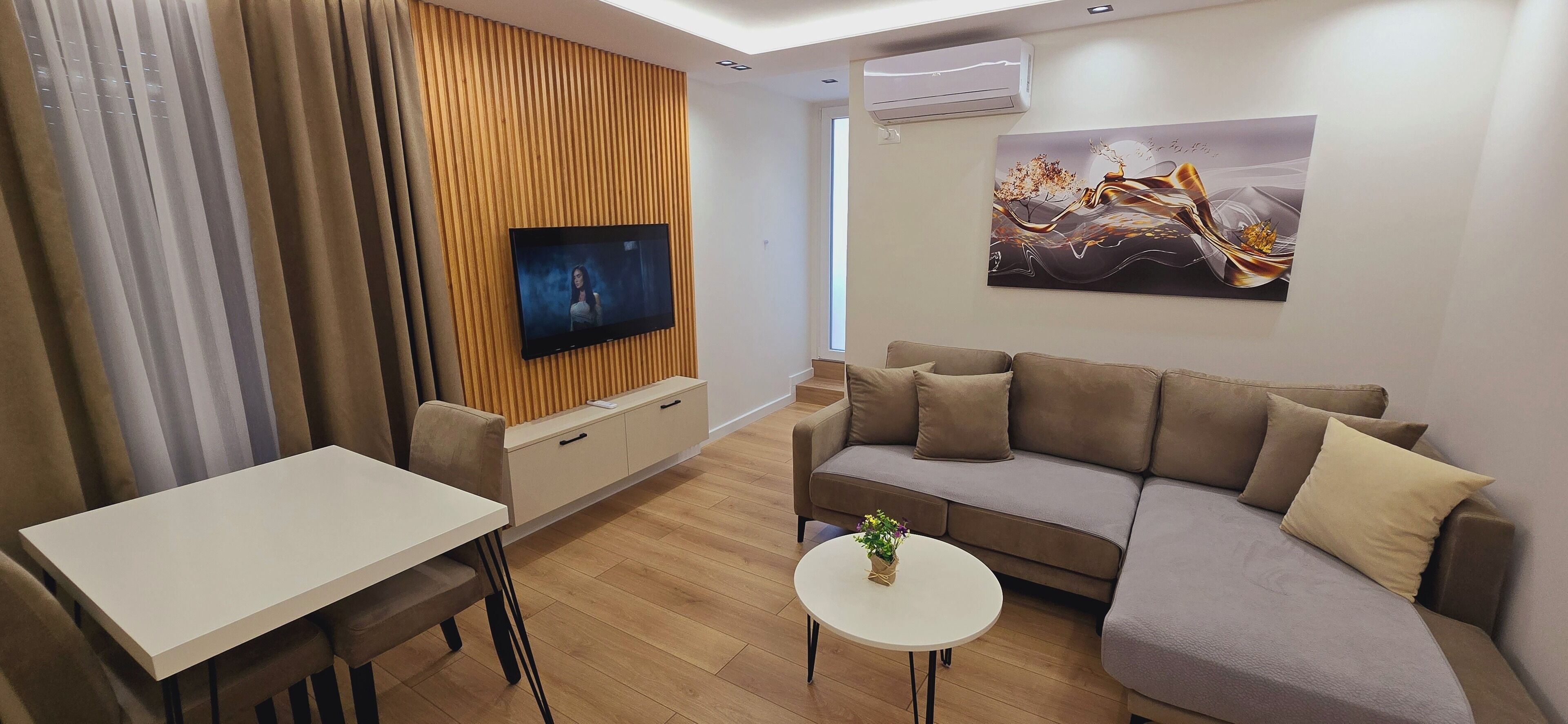 A modern living room with a flat-screen TV mounted on a wooden panel wall, a sofa, a dining table, and a small round table with a plant.