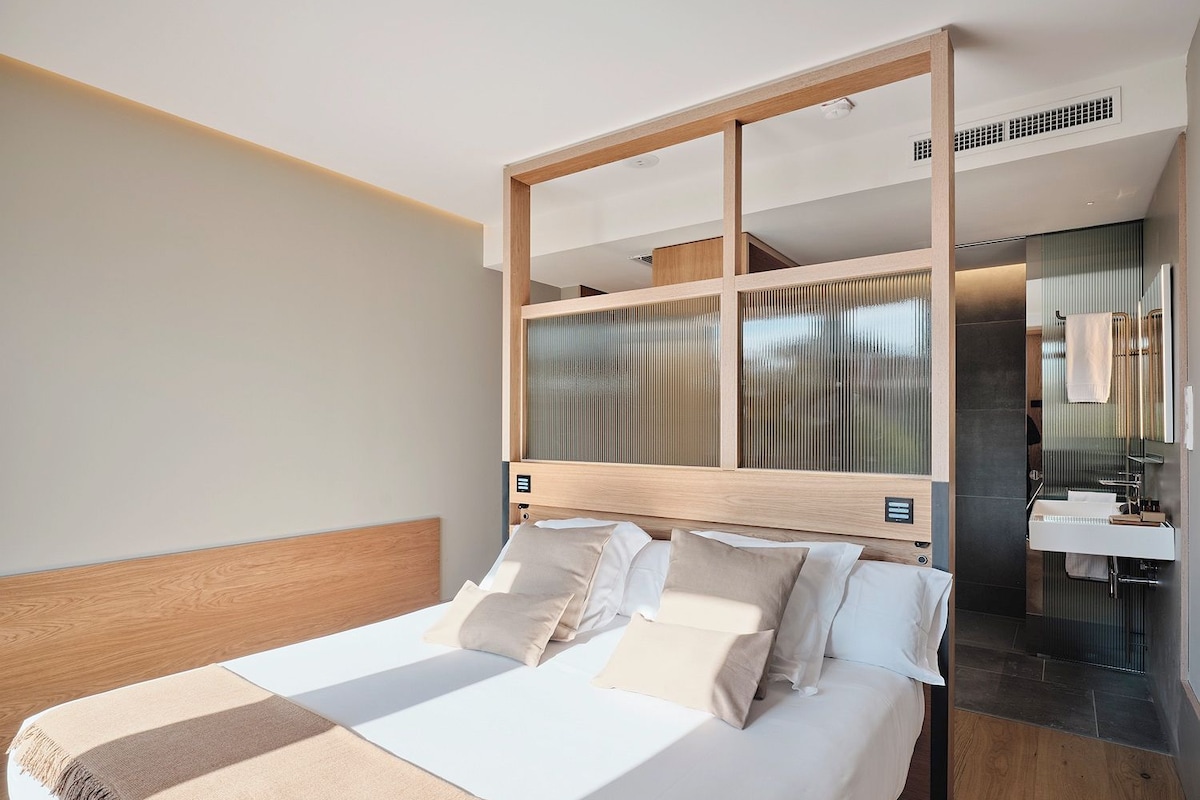 A modern hotel room with a large bed, a wooden headboard, a built-in desk, and a bathroom with a mirror and sink.