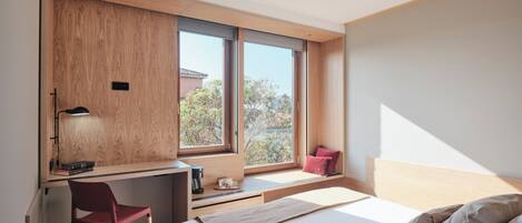 Ein modernes Hotelzimmer mit einem Holztisch, einem roten Sessel, einem Bett mit vielen Kissen und Blick auf Bäume durch große Fenster.