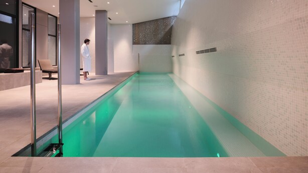 A modern indoor swimming pool with clear water, surrounded by tiled walls and a bench. - Qgat Suites & Events (Sant Cugat del Valles)