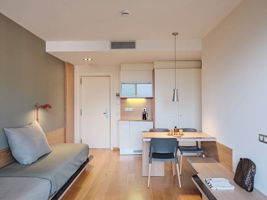 Un espacio habitable compacto con cama, una zona de comedor con sillas, una kitchenette y una pequeña mesa.
