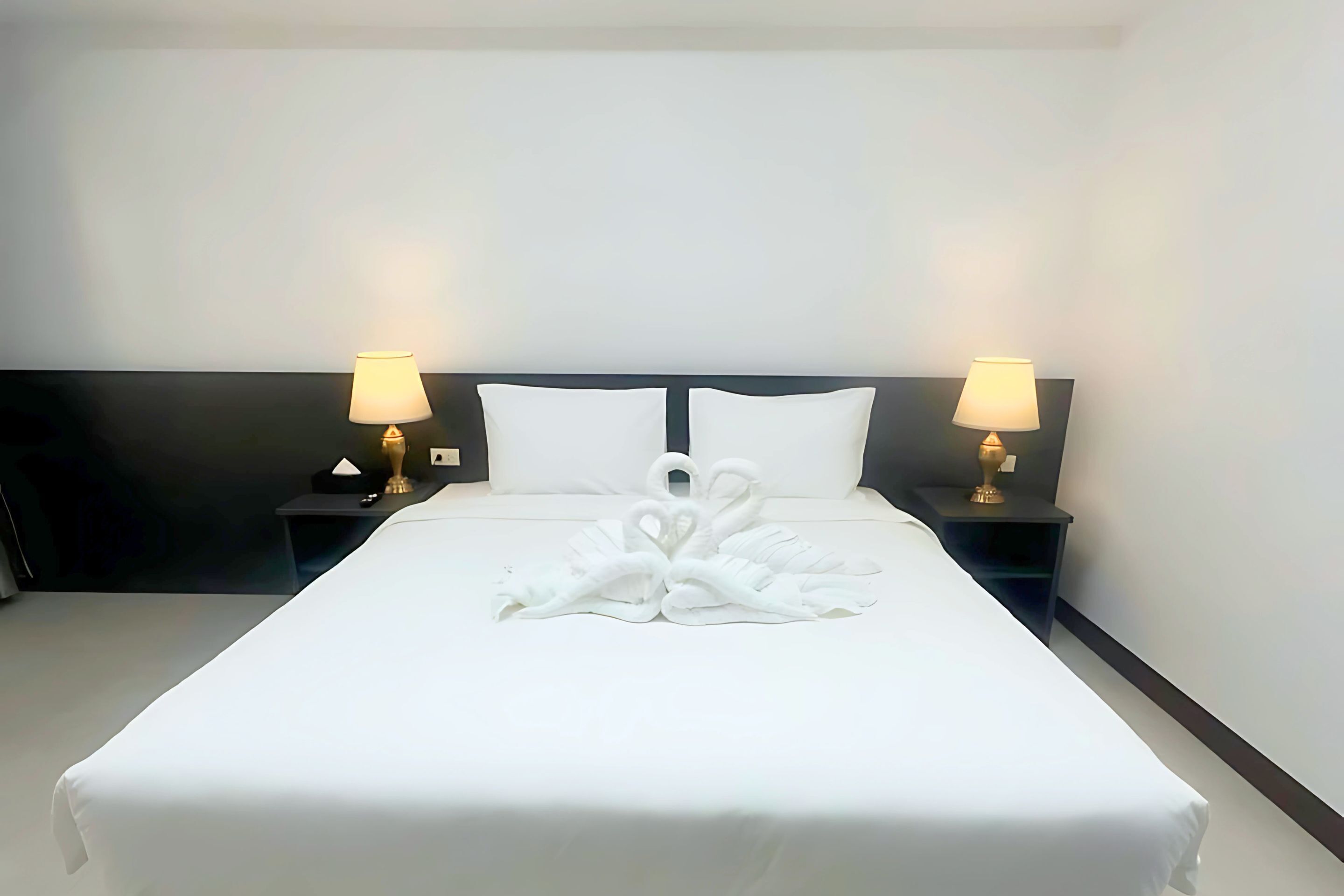 Una cama bien hecha con un centro de toallas con forma de cisne, dos lámparas de noche y un cabecero oscuro.
