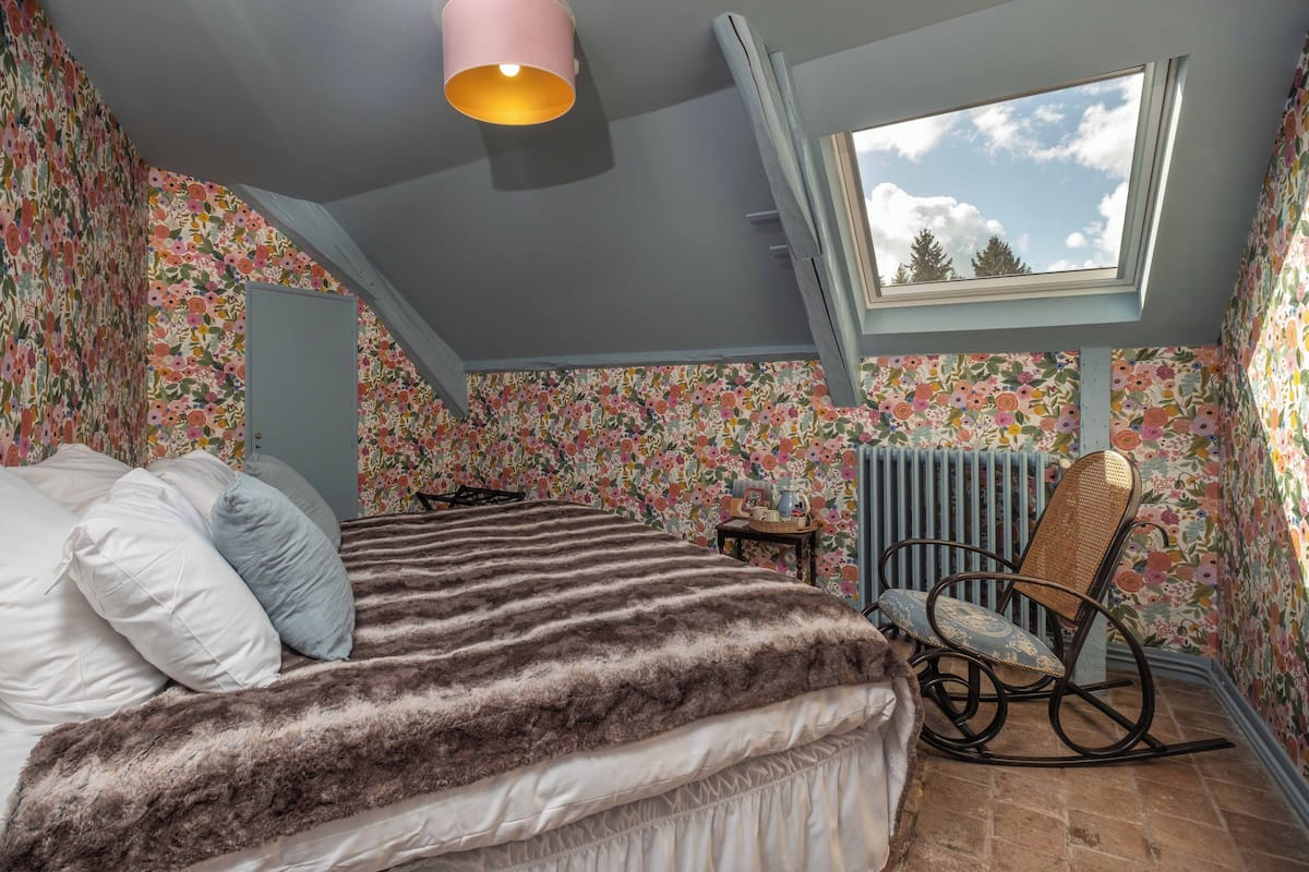 Une chambre à coucher avec un lit, un fauteuil à bascule, une fenêtre et un papier peint à motifs floraux.