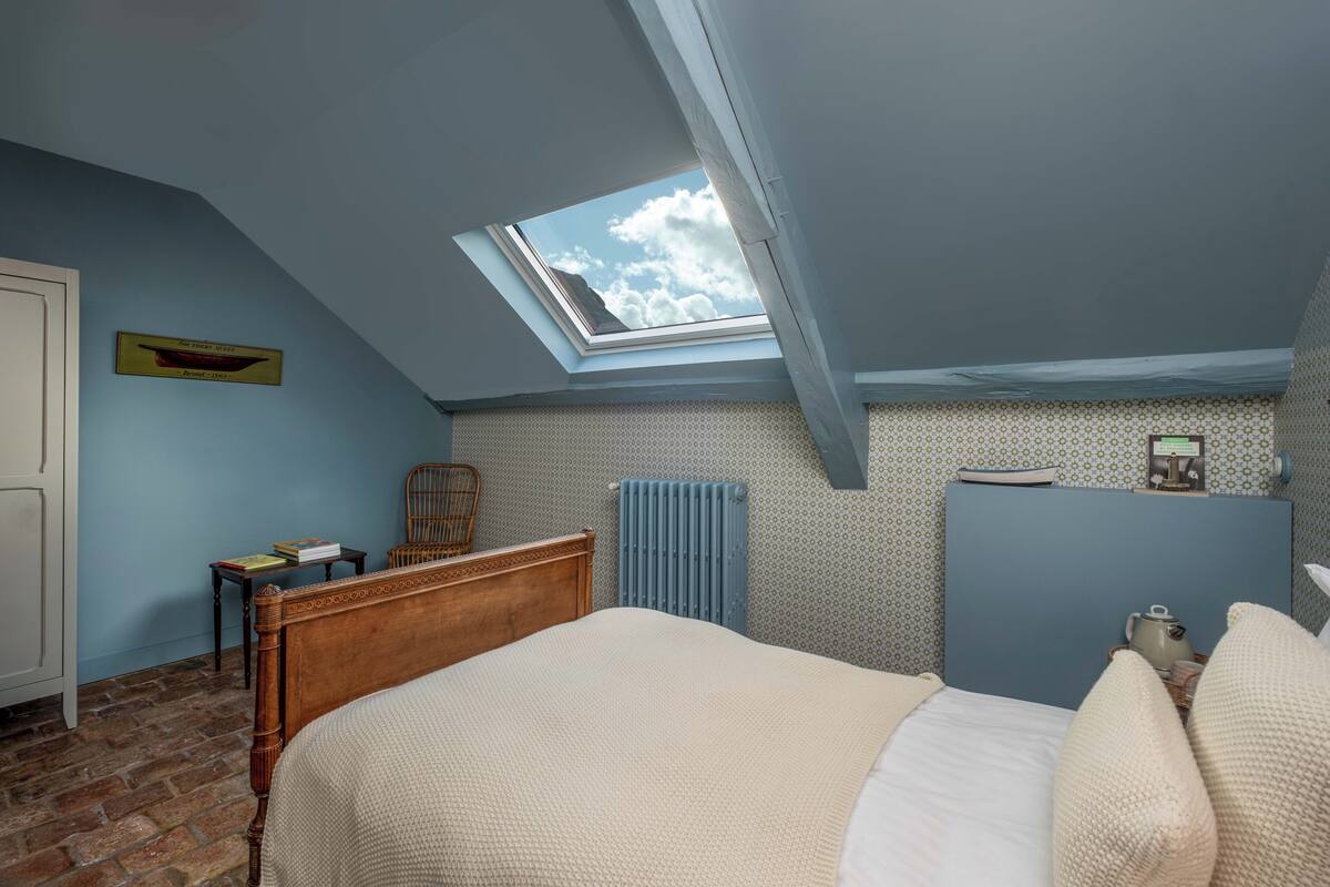 Une chambre avec un plafond en pente, un puits de lumière, un lit en bois, un radiateur, une petite table et une chaise.