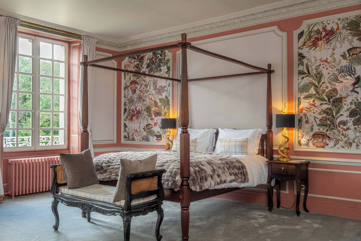 Un lit à baldaquin, une chaise longue, une table de chevet avec une lampe et une grande tapisserie à motifs floraux accrochée au mur.