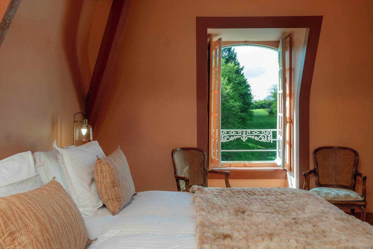 Une chambre avec un grand lit, deux fauteuils en osier et une fenêtre donnant sur un paysage verdoyant.