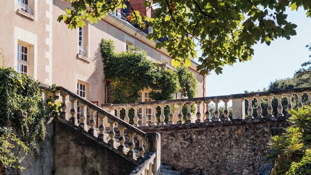 Un escalier en pierre menant à un bâtiment doté d’un balcon, le tout entouré de verdure.