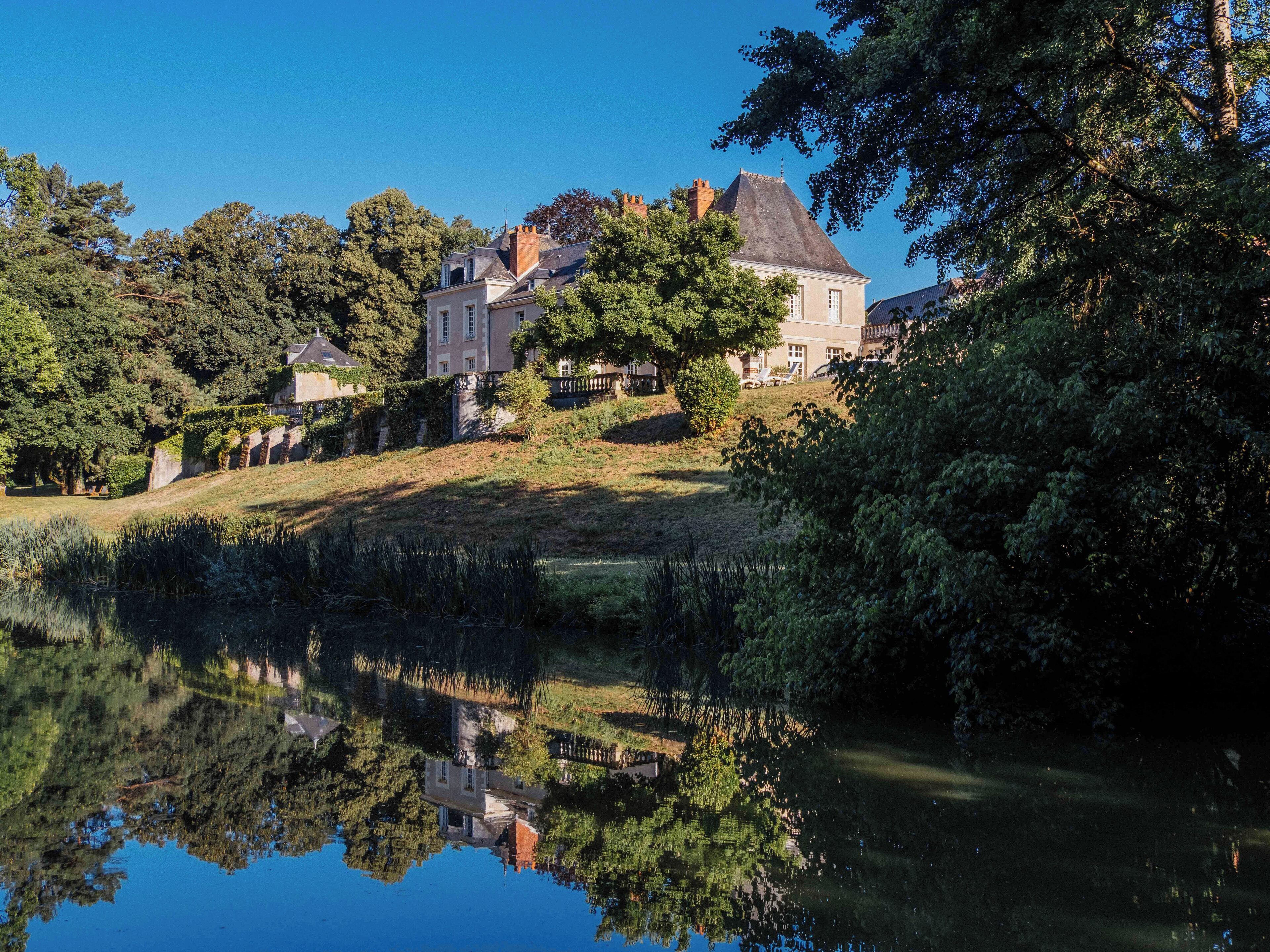 A large, historic building with multiple stories, surrounded by lush greenery and a calm lake reflecting the structure.