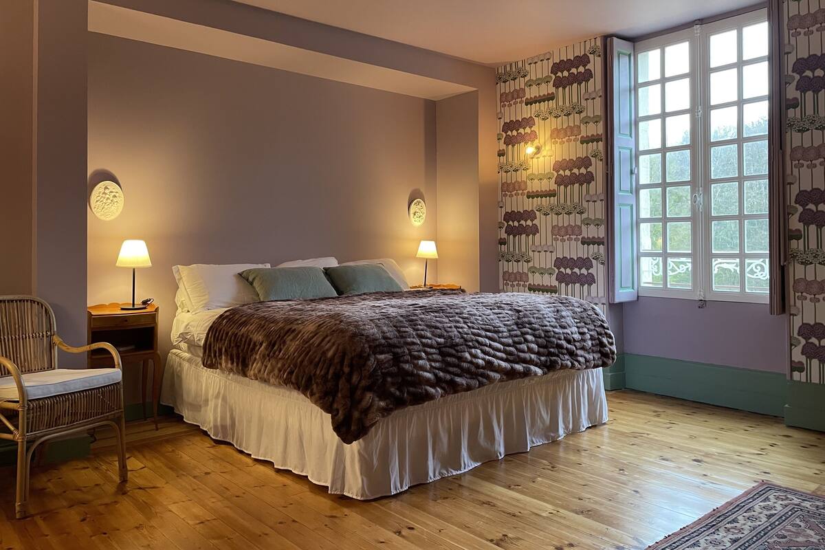 Une chambre à coucher avec un grand lit, deux lampes de chevet, un fauteuil et une fenêtre ornée de rideaux à motifs.