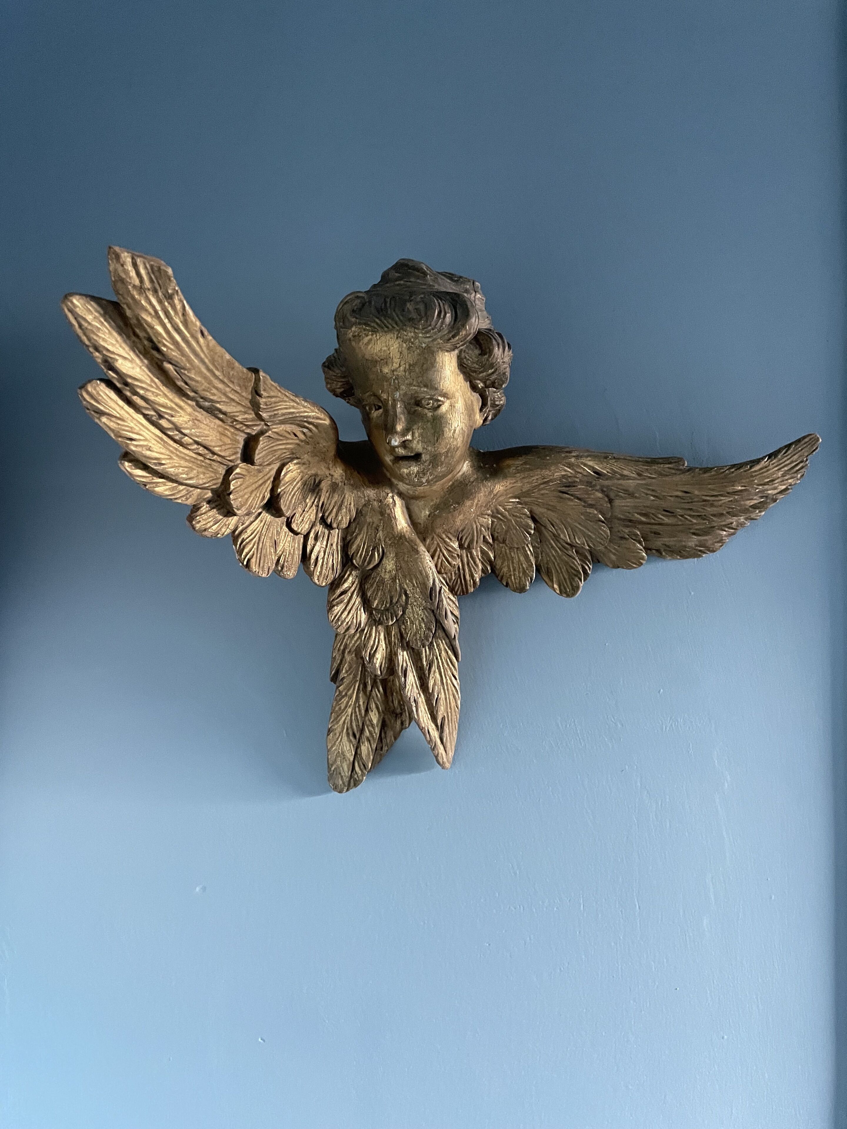 Escultura de anjo em bronze com asas abertas, diante de uma parede azul.