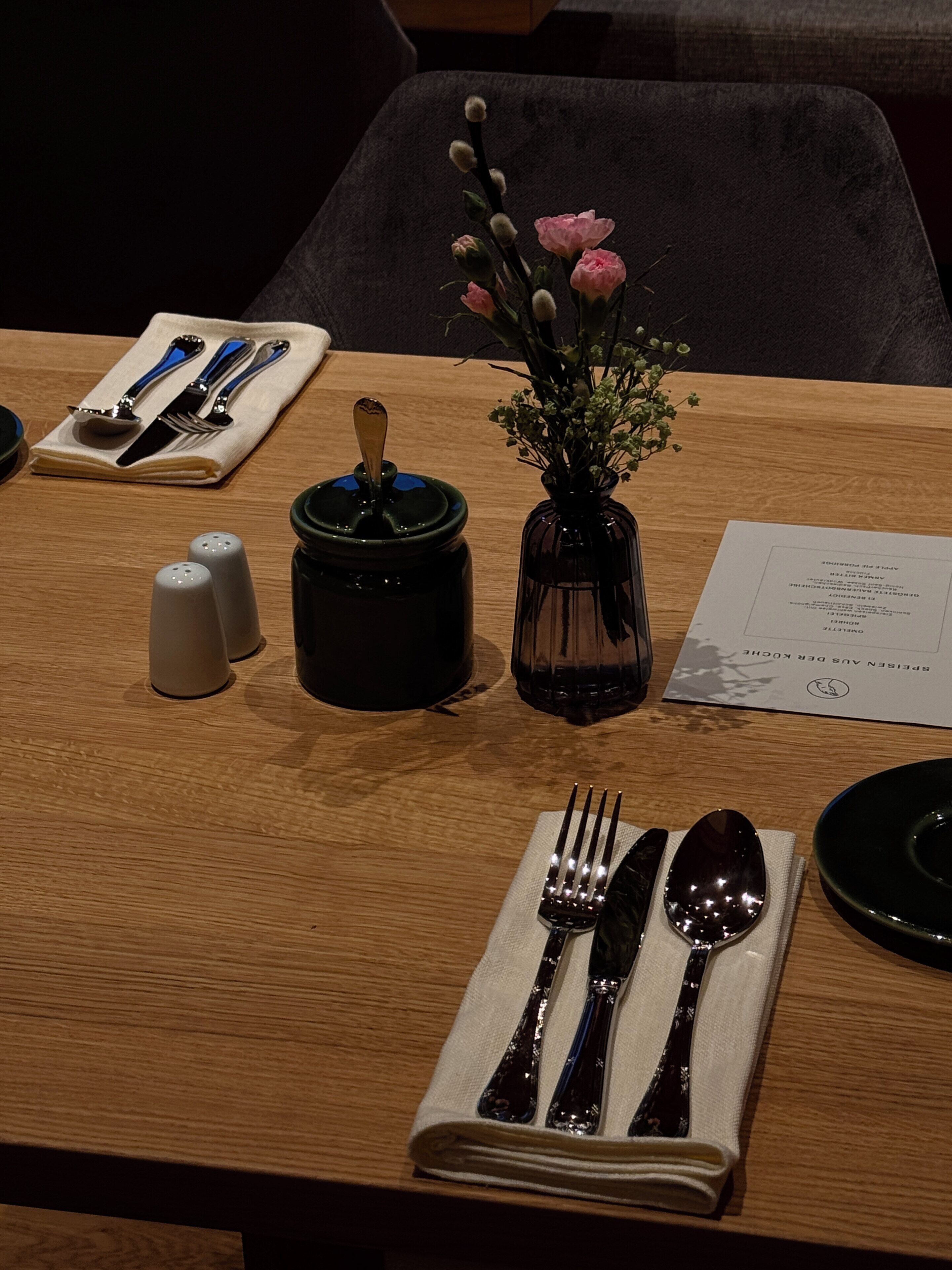Ein ordentlich gedeckter Esstisch mit gefaltetem Servietttuch, Besteck, einer Salzmühlen- und Pfefferstreuer-Kombination, einer kleinen Vase mit Blumen und einer Speisekarte.