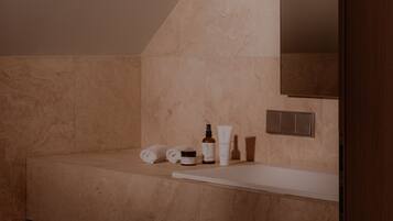 Un baño con pared de azulejos color beige, una encimera blanca y un espejo. Hay artículos de aseo en la encimera.