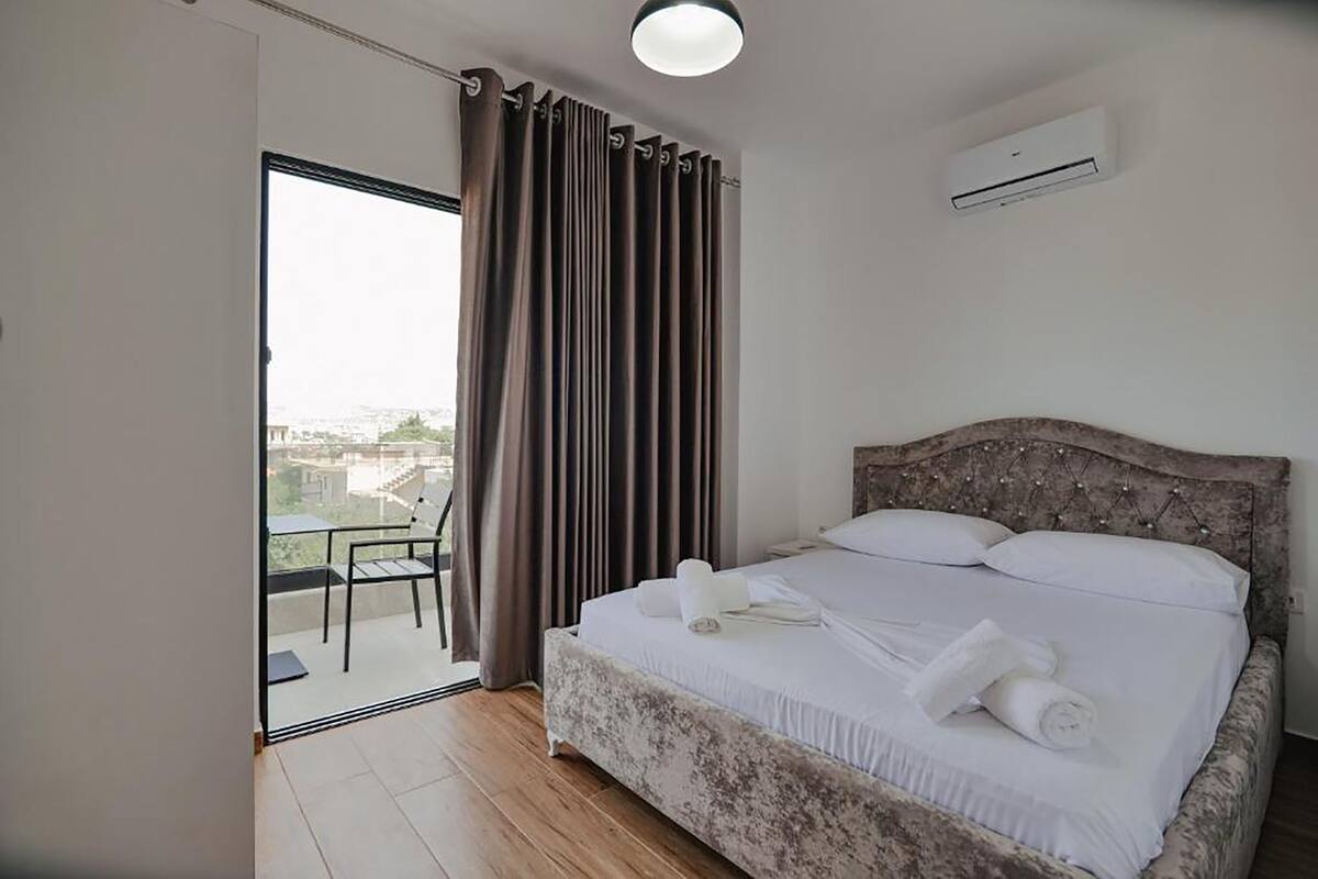 A bedroom with a large bed, a balcony with a chair, and an air conditioning unit on the wall.