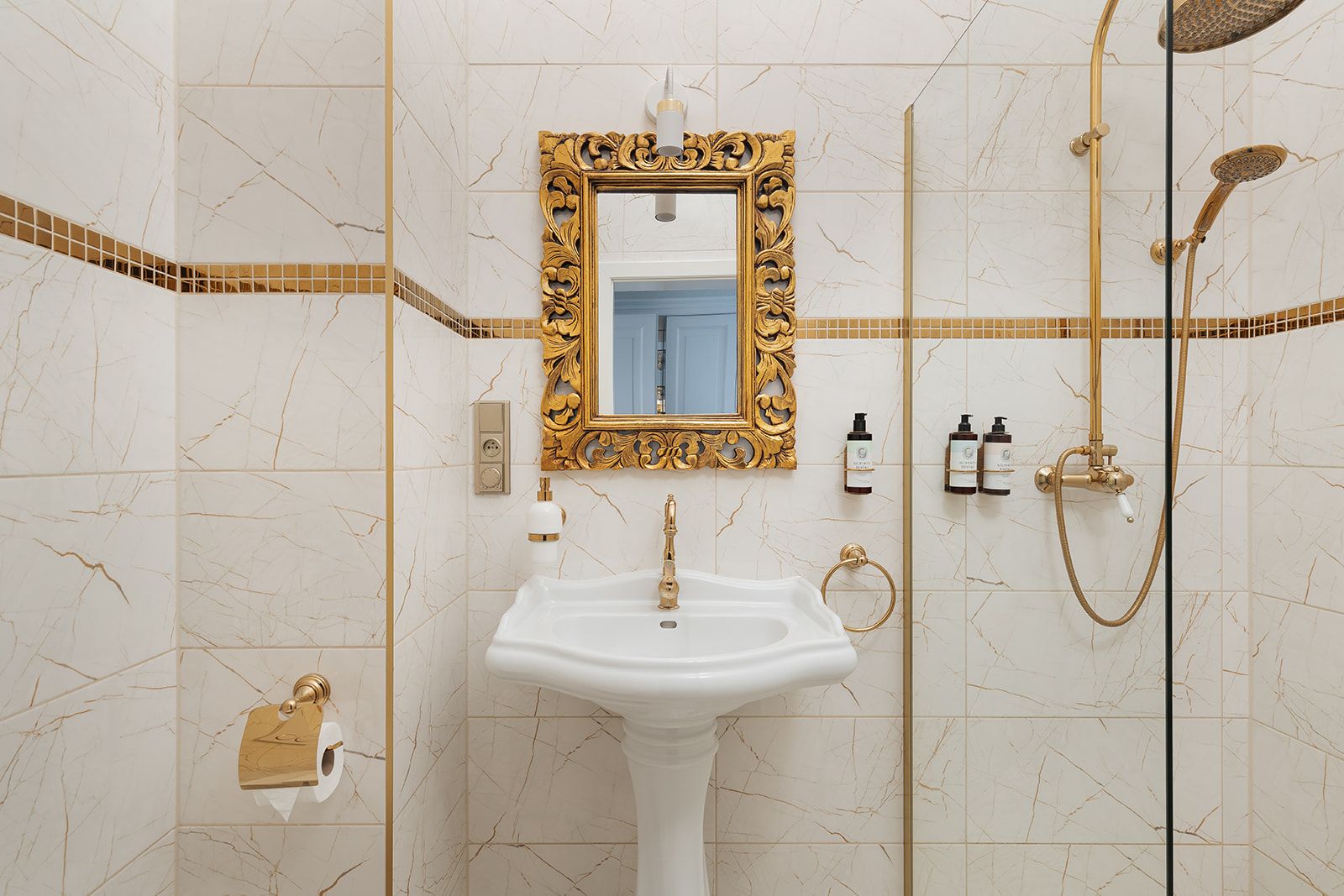 Un baño con lavamanos blanco, espejo con marco dorado y sistema de ducha dorado.