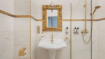 Un baño con lavamanos blanco, espejo con marco dorado y sistema de ducha dorado.