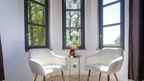 Een kamer met twee witte stoelen, een kleine tafel met een vaas, en grote ramen met uitzicht op bomen.