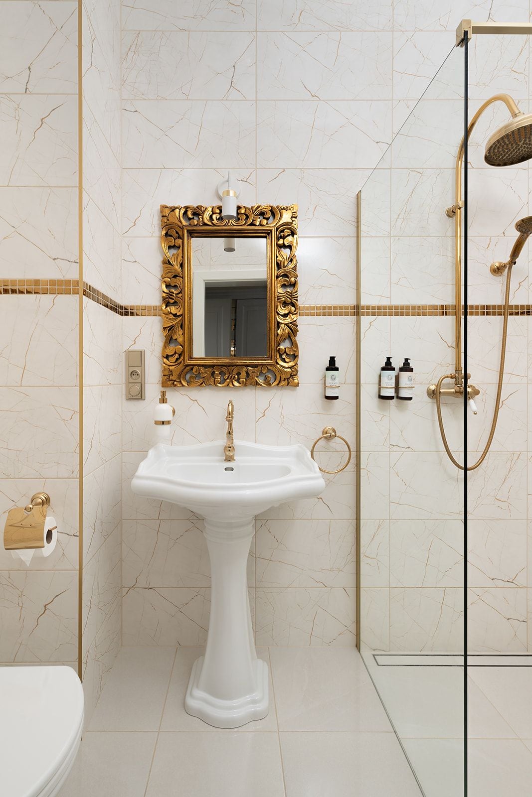 Casa de banho com lavatório pedestal branco, espelho com moldura dourada, chuveiro dourado e sanita.